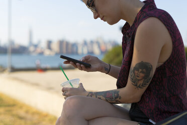 USA, New York City, Brooklyn, tätowierte Frau sitzt auf einer Bank und schaut auf ihr Smartphone - GIOF000687