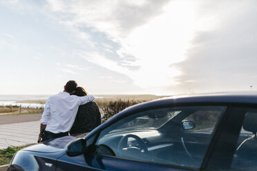 Spain, Cubelles, back view of a young couple sitting on the hood of the car watching the sea at sunset - JRFF000351