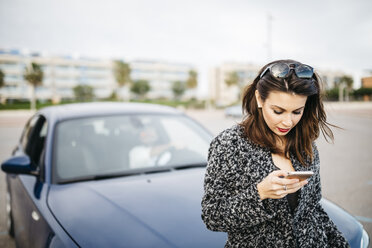 Spanien, Cubelles, junge Frau, die auf der Motorhaube des Autos sitzt und auf ihr Smartphone schaut - JRFF000352