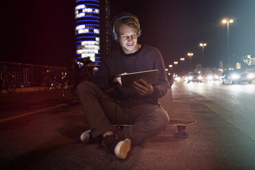 Deutschland, München, Mann mit Kopfhörern, der auf seinem Skateboard sitzt und nachts ein digitales Tablet benutzt - RBF004073