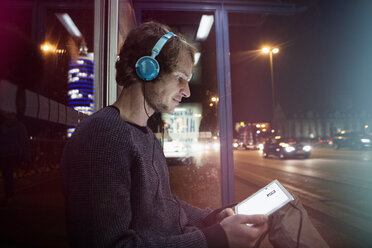 Deutschland, München, Mann mit Kopfhörern, der an der Bushaltestelle sitzt und nachts ein digitales Tablet benutzt - RBF004074