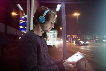 Germany, Munich, man with headphones sitting at bus stop using digital tablet at night - RBF004074