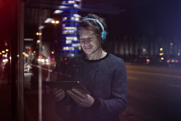 Germany, Munich, man with headphones standing at bus stop using digital tablet at night - RBF004076