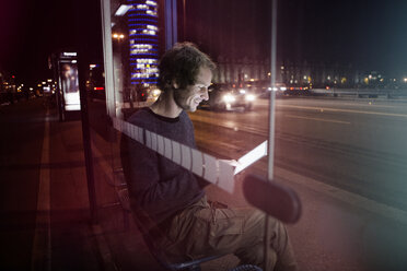 Germany, Munich, man with headphones sitting at bus stop using digital tablet at night - RBF004078