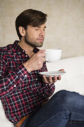 Porträt eines Mannes mit geschlossenen Augen, der auf einer Couch sitzt und eine Tasse Kaffee hält - SHKF000467