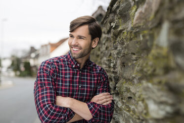 Portrait of smiling man wearing checked shirt leaning against stone wall - SHKF000469