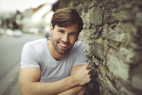 Portrait of smiling man wearing t-shirt leaning against stone wall - SHKF000470