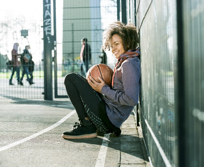 Junge Frau mit Basketball auf einem Spielfeld hockend - MADF000777