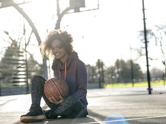 Portrait of smiling young woman with basketball sitting on a playing field - MADF000786