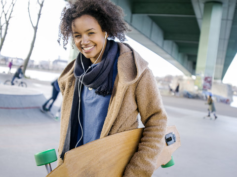 Lächelnde junge Frau mit Longboard, die mit Kopfhörern Musik hört, lizenzfreies Stockfoto