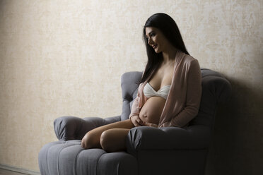 Lächelnde schwangere Frau in Unterwäsche auf einem Sessel sitzend - SHKF000481