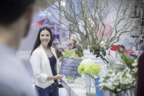 Lächelnde junge Frau in einem Blumenladen zeigt einem Mann ein Schild, lizenzfreies Stockfoto