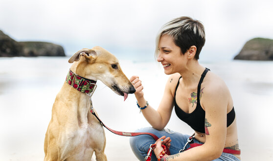 Spanien, Llanes, lächelnde junge Frau mit ihrem Windhund am Strand - MGOF001298