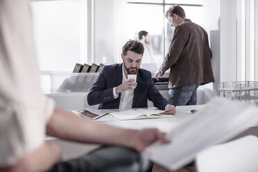 Mann sitzt am Schreibtisch und trinkt Kaffee in einem geschäftigen Büro - ZEF008138