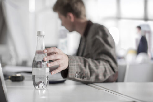 Mann im Büro am Schreibtisch greift nach einer Wasserflasche - ZEF008161