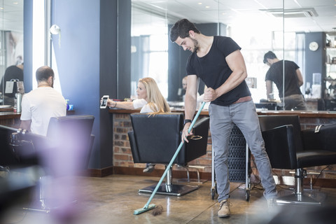 Friseurin kehrt Boden im Salon mit Kunden im Hintergrund, lizenzfreies Stockfoto