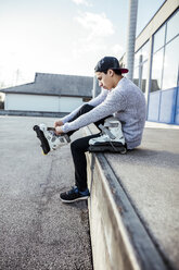 Junger Mann sitzt auf einer Rampe und zieht Inline-Skates an - DAWF000489