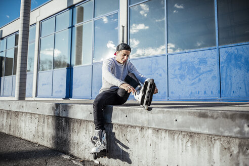 Junger Mann sitzt auf einer Rampe und zieht Inline-Skates an - DAWF000490