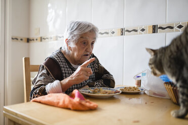 Senior woman sitting at dining table scolding her cat - RAEF000819