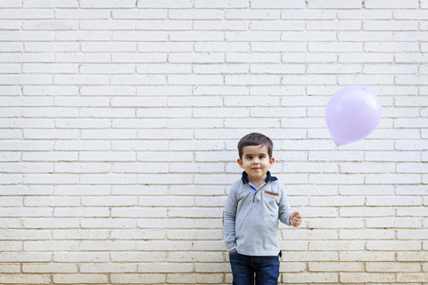 Porträt eines Kleinkindes, das vor einer weißen Wand steht und einen Luftballon hält, lizenzfreies Stockfoto