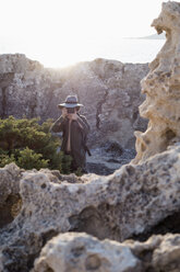 Spain, Ibiza, photographer standing in rocks wearing floppy hat taking a picture - NDF000569