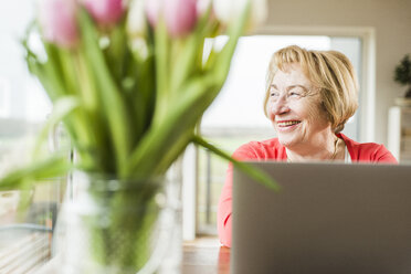 Lächelnde ältere Frau sitzt am Tisch mit Laptop - UUF006427