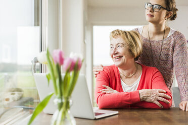 Lächelnde junge Frau mit älterer Frau am Tisch mit Laptop und Blick aus dem Fenster - UUF006431