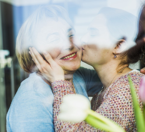 Young woman kissing senior woman behind windowpane stock photo