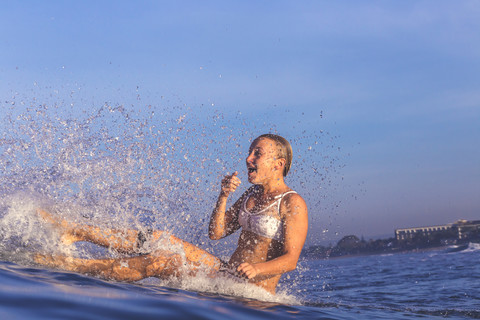 Indonesien, Bali, Frau auf ihrem Surfbrett sitzend und mit Wasser spritzend, lizenzfreies Stockfoto