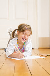 Portrait of laughing little girl lying on wooden floor with crayons and sheet of paper - LVF004510
