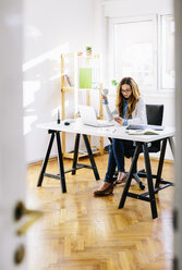 Junge Frau mit einer Tasse Kaffee und einem Smartphone am Schreibtisch in ihrem Heimbüro sitzend - AKNF000036