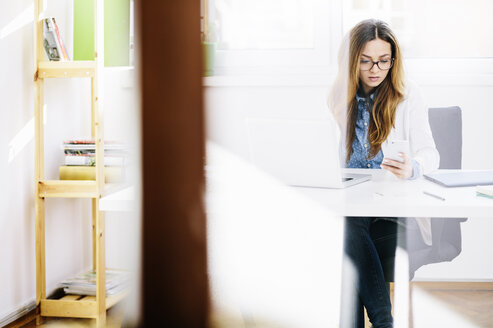 Young woman sitting at desk in her home office looking at smartphone - AKNF000037