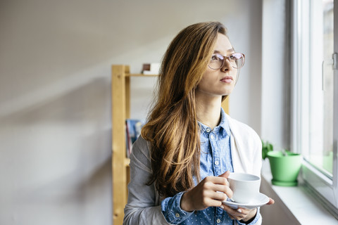 Junge Frau, die sich mit einer Tasse Kaffee ausruht und durch das Fenster in ihr Büro schaut, lizenzfreies Stockfoto