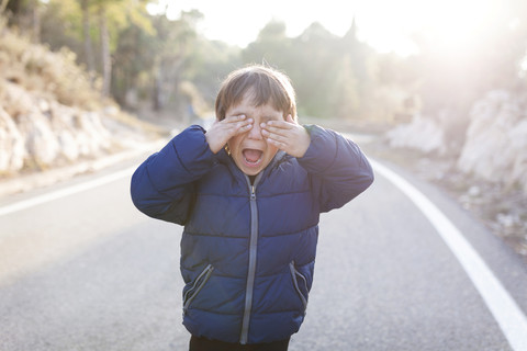 Spanien, Siurana, schreiender kleiner Junge, der auf der Fahrbahn einer leeren Landstraße steht und sich die Augen mit den Händen bedeckt, lizenzfreies Stockfoto