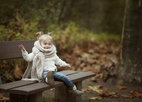 Blondes kleines Mädchen mit großem Schal sitzt auf einer Bank im Herbstwald, lizenzfreies Stockfoto