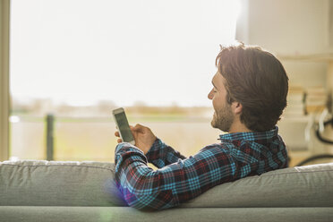 Lächelnder junger Mann lehnt an der Rückenlehne der Couch und schaut auf sein Smartphone - UUF006466