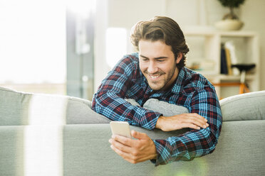 Porträt eines lächelnden jungen Mannes, der sich an die Rückenlehne der Couch lehnt und auf sein Smartphone schaut - UUF006468