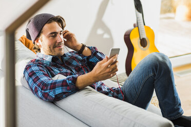 Porträt eines jungen Mannes, der auf der Couch sitzt und mit Kopfhörern und Smartphone Musik hört - UUF006472