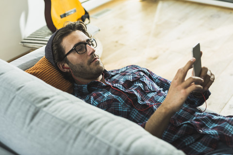 Junger Mann liegt auf der Couch und hört Musik mit Kopfhörern und Smartphone, lizenzfreies Stockfoto