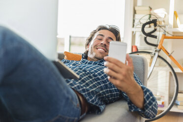 Junger Mann liegt auf der Couch mit Laptop und schaut auf sein Smartphone - UUF006486