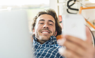 Porträt eines lächelnden jungen Mannes, der mit einem Laptop auf der Couch liegt und auf sein Smartphone schaut - UUF006489