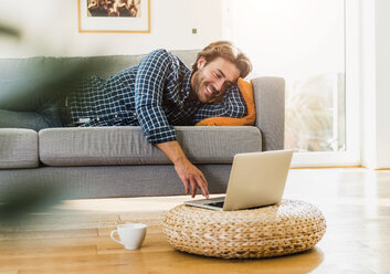 Junger Mann liegt zu Hause auf der Couch und schaut auf seinen Laptop - UUF006490