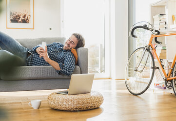 Young man lying on the couch at home looking at laptop - UUF006491