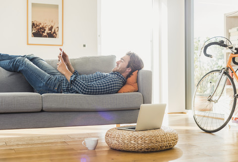 Junger Mann liegt zu Hause auf der Couch und schaut auf ein digitales Tablet, lizenzfreies Stockfoto