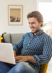 Porträt eines lächelnden jungen Mannes, der im Wohnzimmer sitzt und einen Laptop benutzt - UUF006495