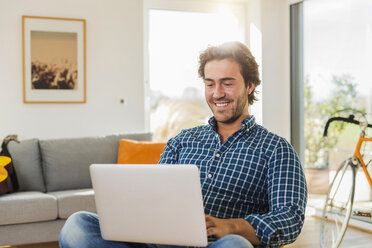 Porträt eines lächelnden jungen Mannes, der im Wohnzimmer sitzt und einen Laptop benutzt - UUF006496