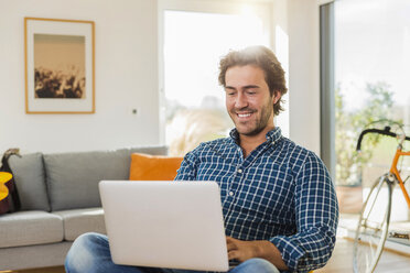 Portrait of smiling young man sitting in the living room using laptop - UUF006496