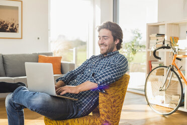 Porträt eines lächelnden jungen Mannes, der auf einem Sessel im Wohnzimmer sitzt und einen Laptop benutzt - UUF006498