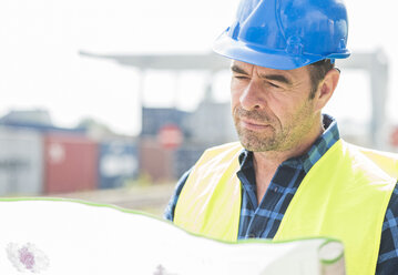 Mann mit Schutzhelm beim Lesen eines Dokuments im Containerhafen - UUF006523