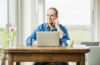 Businessman at home with laptop at wooden table thinking - UUF006541
